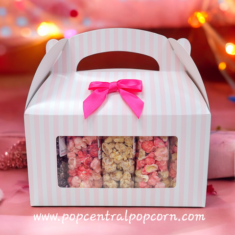 Striped popcorn box with pink bow and window displaying popcorn, on a blurred background.